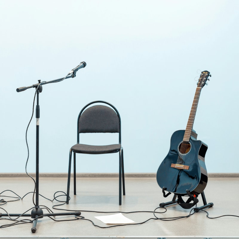 A microphone stand with a microphone and a sheet of paper are in the foreground, with a chair and an acoustic guitar on a stand in the background against a light blue wall.