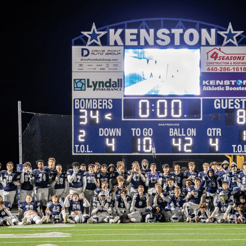 A football team poses in front of a brightly lit scoreboard at night.