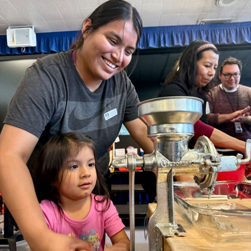 A young girl and a woman smile while operating a manual grinder in the foreground, with other people visible in the blurred background.