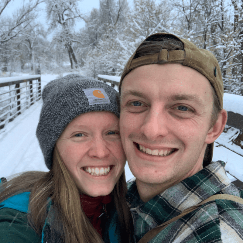 A smiling couple in winter hats and jackets pose for a selfie on a snow-covered bridge with trees in the background.