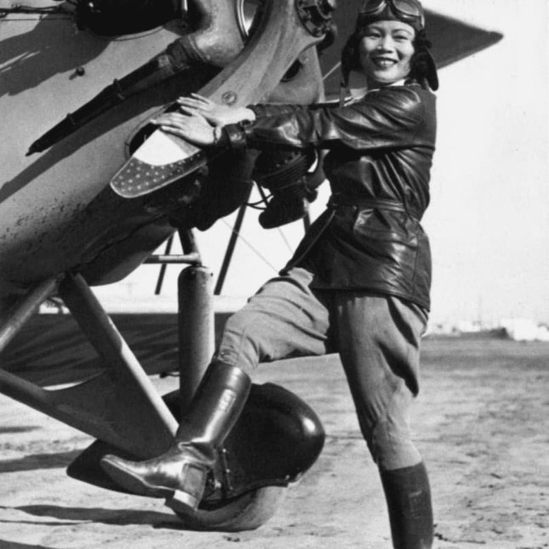 A smiling woman in aviator gear poses with her hand on the propeller of a biplane against a bright, open background.