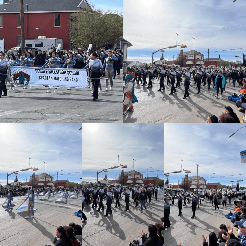 A marching band with a banner is parading down a street lined with spectators and buildings under a bright sky.