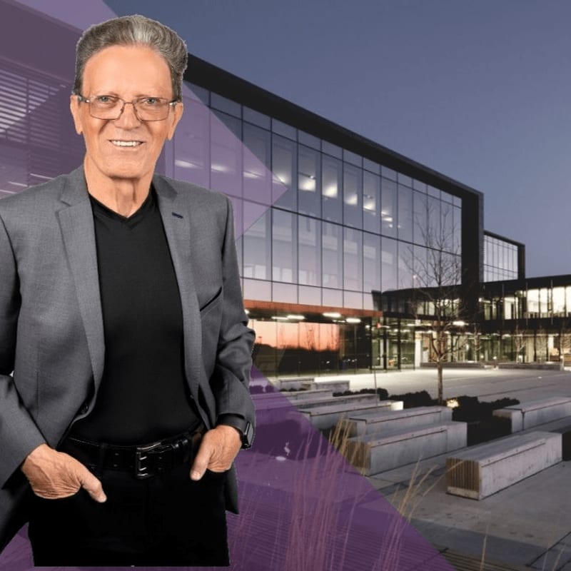 A smiling older man in a suit stands in the foreground with a modern glass building in the background.