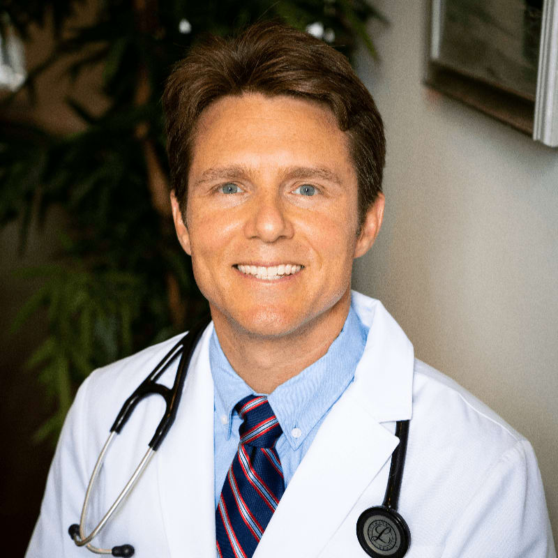 A smiling man in a white lab coat and stethoscope stands in front of a blurred background with a plant and a framed picture.