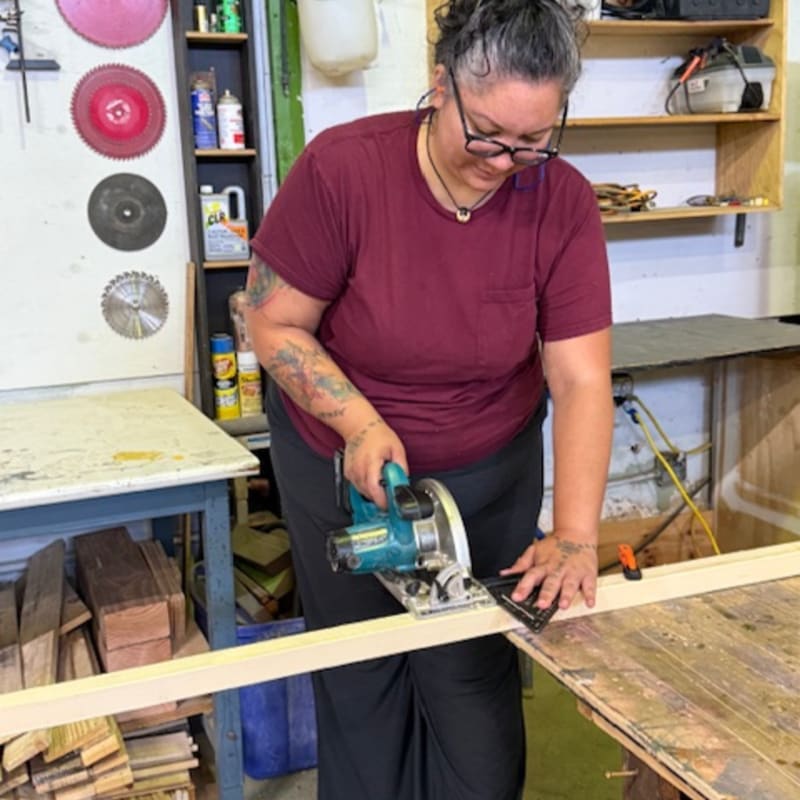 A woman in a maroon shirt uses a circular saw on a wooden plank in the foreground, while shelves filled with tools and supplies are visible in the background.
