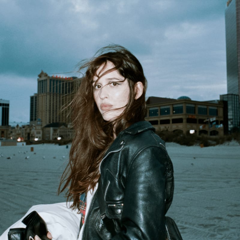 A woman in a leather jacket stands on a sandy beach with buildings in the background under a cloudy sky.
