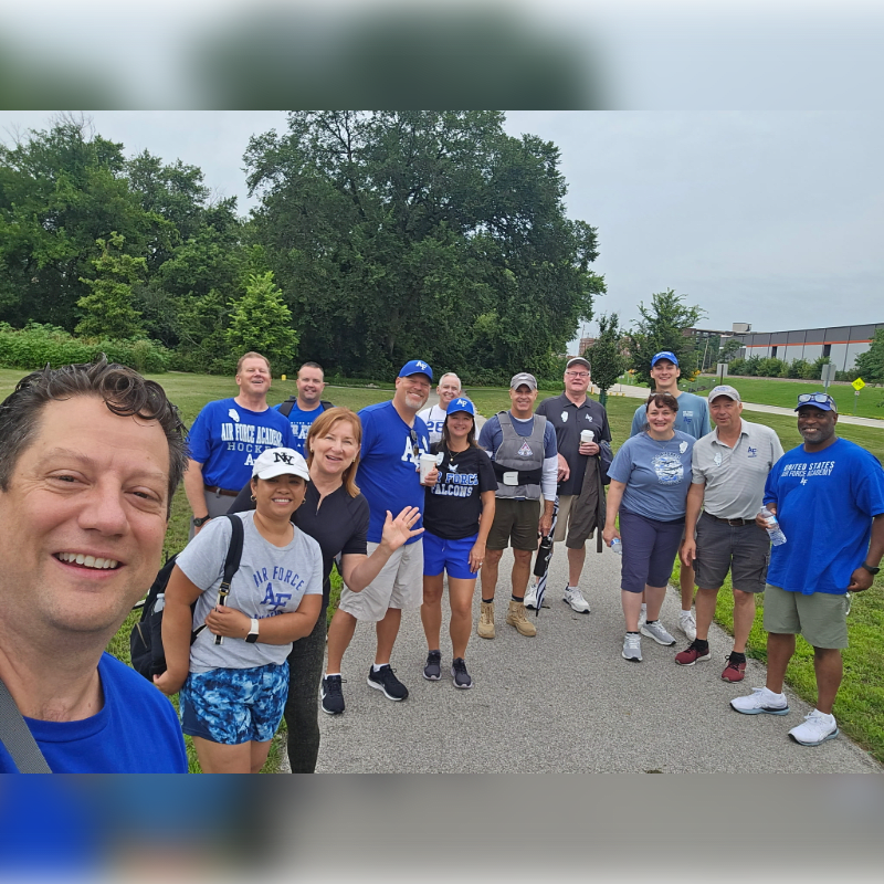 A group of people in blue shirts pose for a selfie on a paved path with trees and buildings in the background.