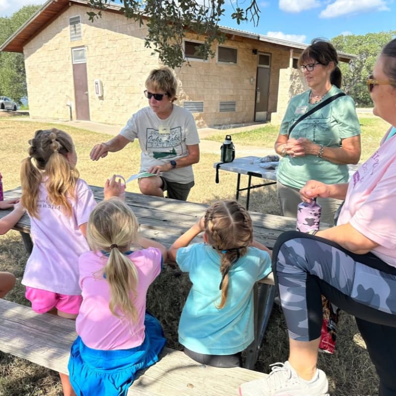 Children in the foreground sit at a picnic table listening to an adult, while a building and trees form the background.