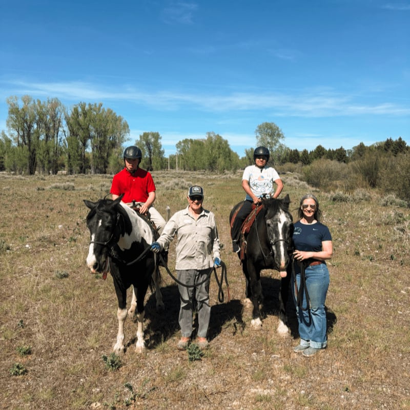 Four people on horseback stand on a dry, grassy path with trees and a clear blue sky in the background.