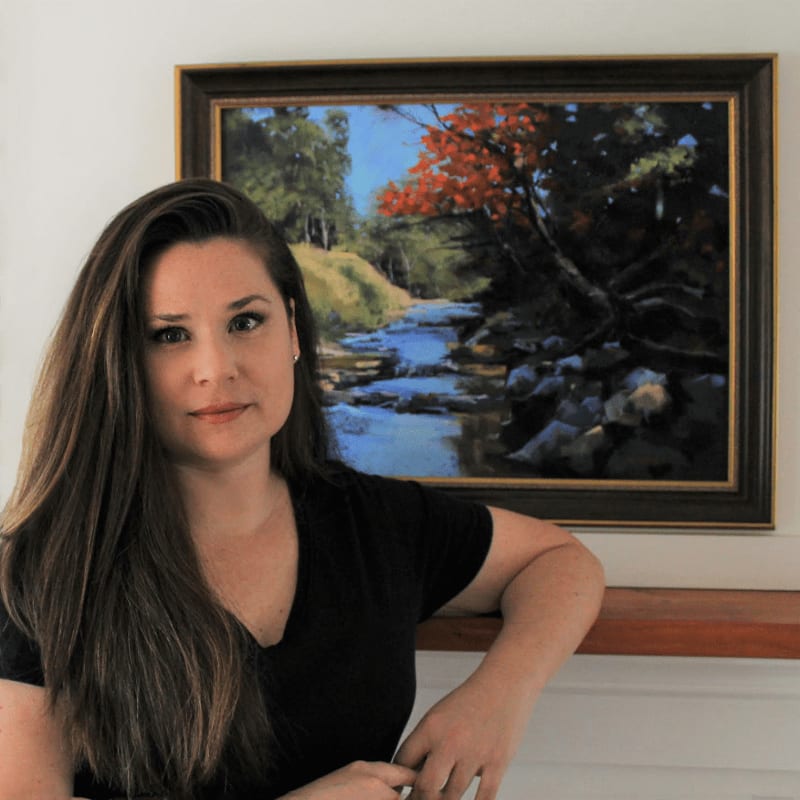 A woman with long brown hair leans on a wooden surface in front of a framed painting of a vibrant autumn landscape with a flowing stream.