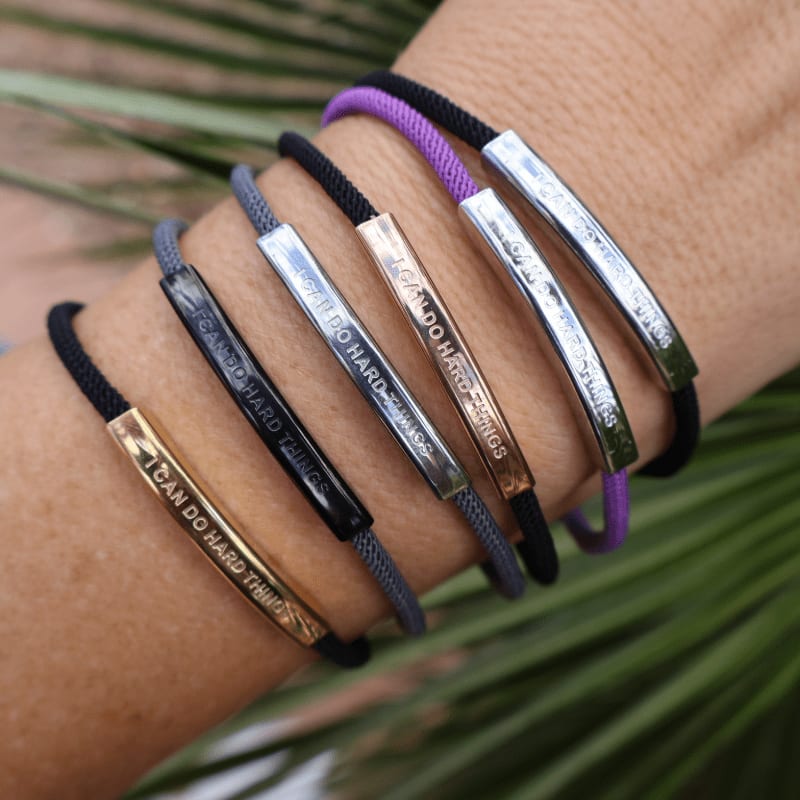 Several bracelets with inspirational messages are worn on a wrist, with blurred green foliage in the background.