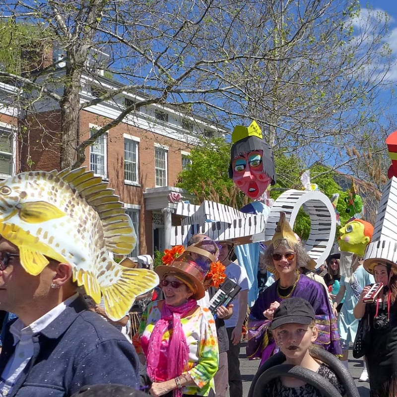 People in elaborate, whimsical hats, including a large fish and musical notes, parade down a street lined with brick buildings under a bright sky.