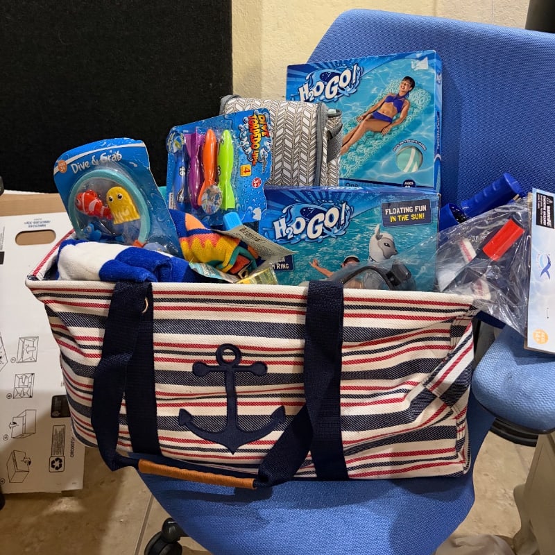 A striped tote bag filled with pool toys sits on a blue chair, with boxes of pool floats and other items in the background.