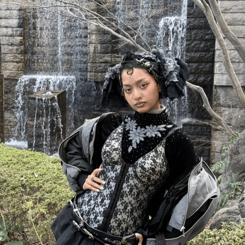 A person with elaborate hair and a patterned outfit stands in front of a rocky waterfall and lush greenery.
