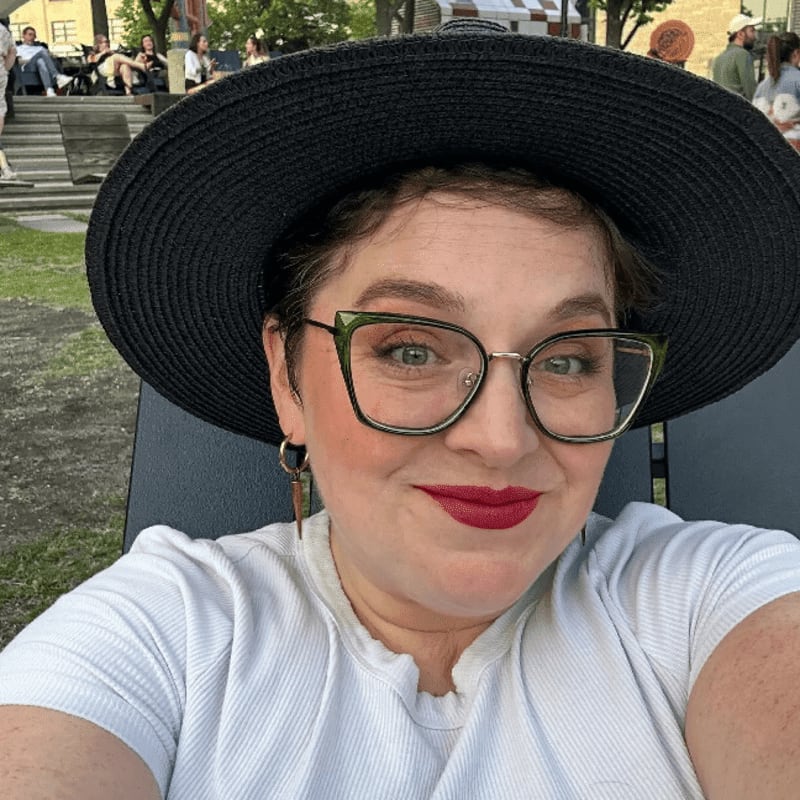 A woman in a black hat and glasses smiles at the camera in the foreground, while people relax on steps in a park in the background.