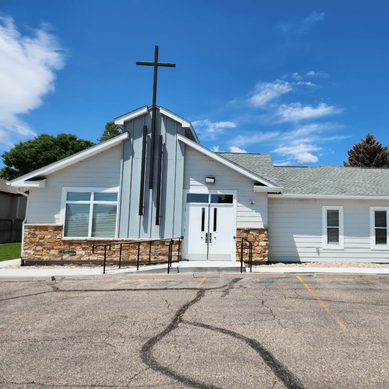 A light blue church with a stone base and a cross on top stands under a bright blue sky with fluffy clouds, with a cracked asphalt parking lot in the foreground.