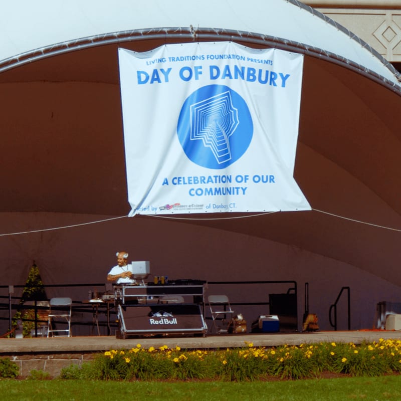 A DJ with a fox mask stands behind a Red Bull-branded setup on a stage with a "Day of Danbury" banner, set against a backdrop of a large white tent and a building.
