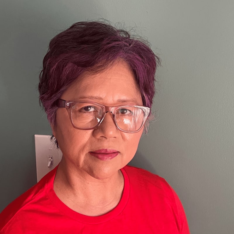 A woman with purple-tinted hair and glasses wears a red shirt against a muted green wall with a light switch.