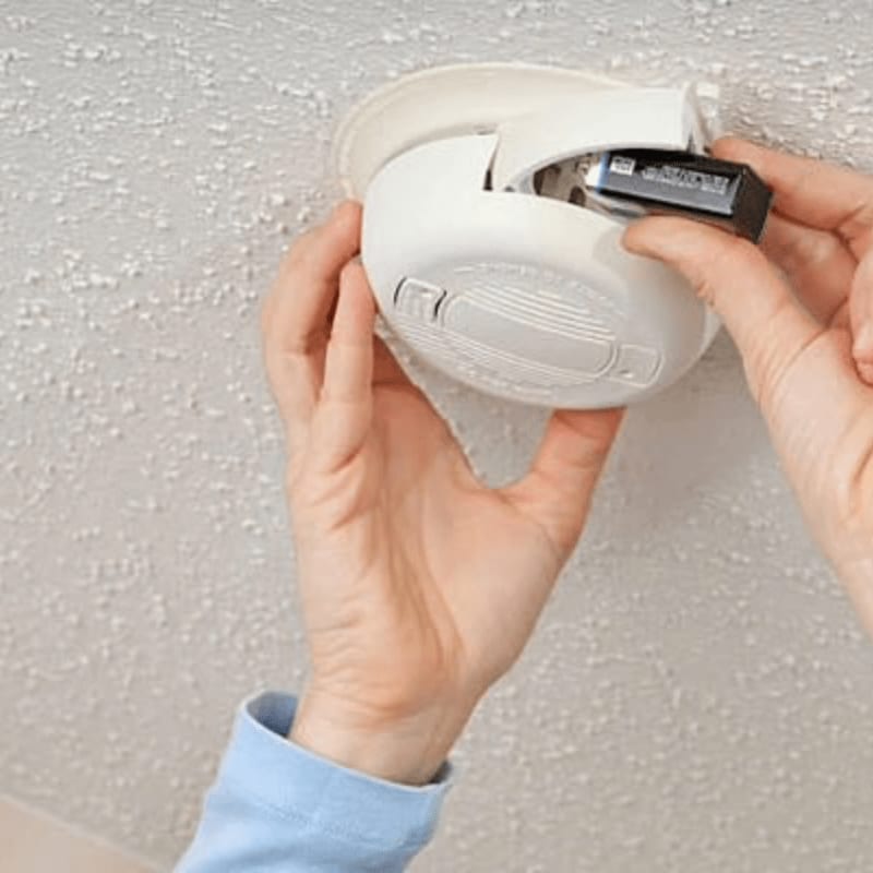 Hands insert a battery into a white smoke detector mounted on a textured ceiling.