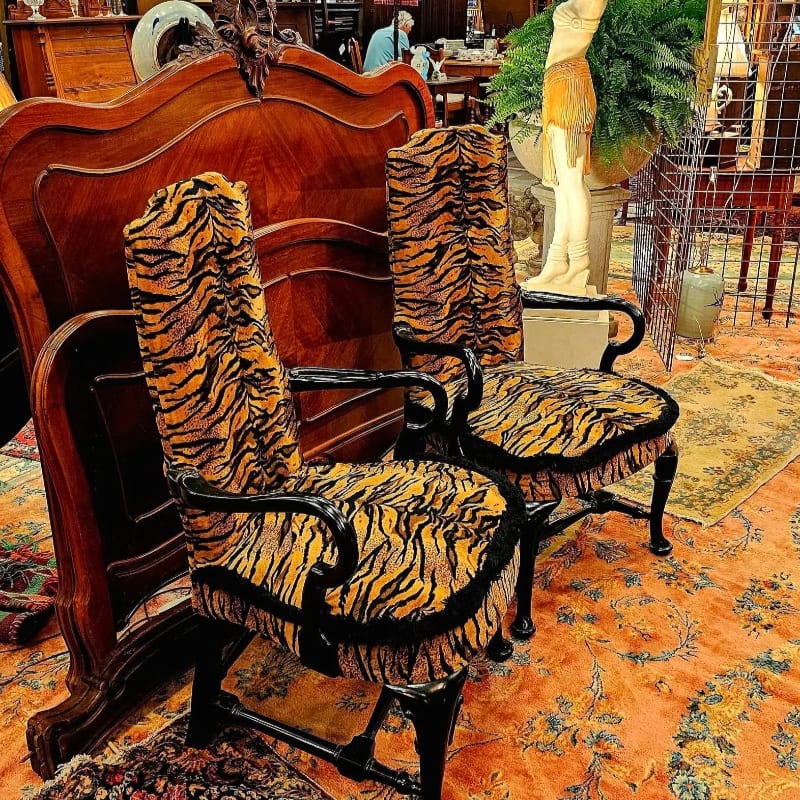 Two tiger-striped chairs with ornate wooden frames sit on a patterned rug in front of a large, dark wooden headboard, with a statue and greenery visible in the blurred background.