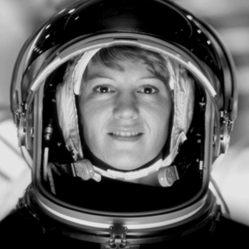 A smiling astronaut in a helmet looks forward, with a blurred, dark background.