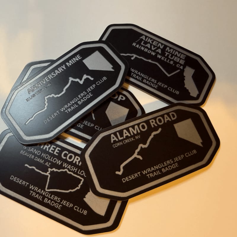 A pile of black and silver trail badges, each featuring a map and location, rests on a light-colored surface.