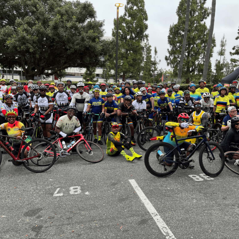A large group of cyclists in yellow shirts pose with their bikes in the foreground, while trees and buildings form the background.