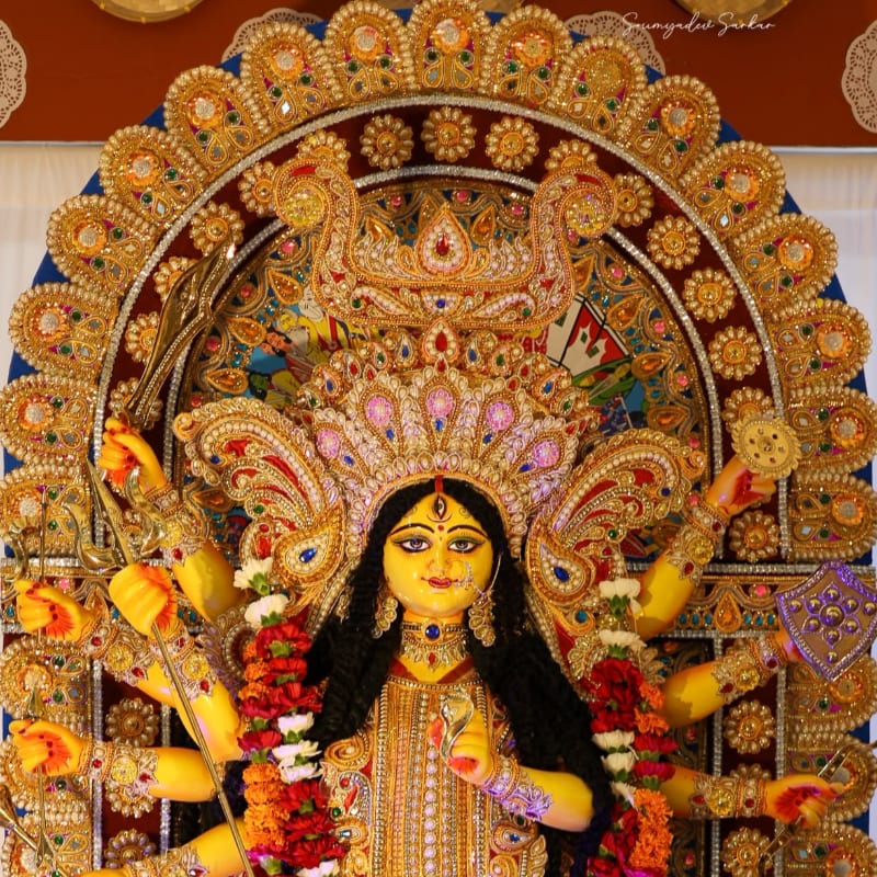 A brightly adorned idol of Goddess Durga with multiple arms is the central focus, set against an ornate, golden archway.