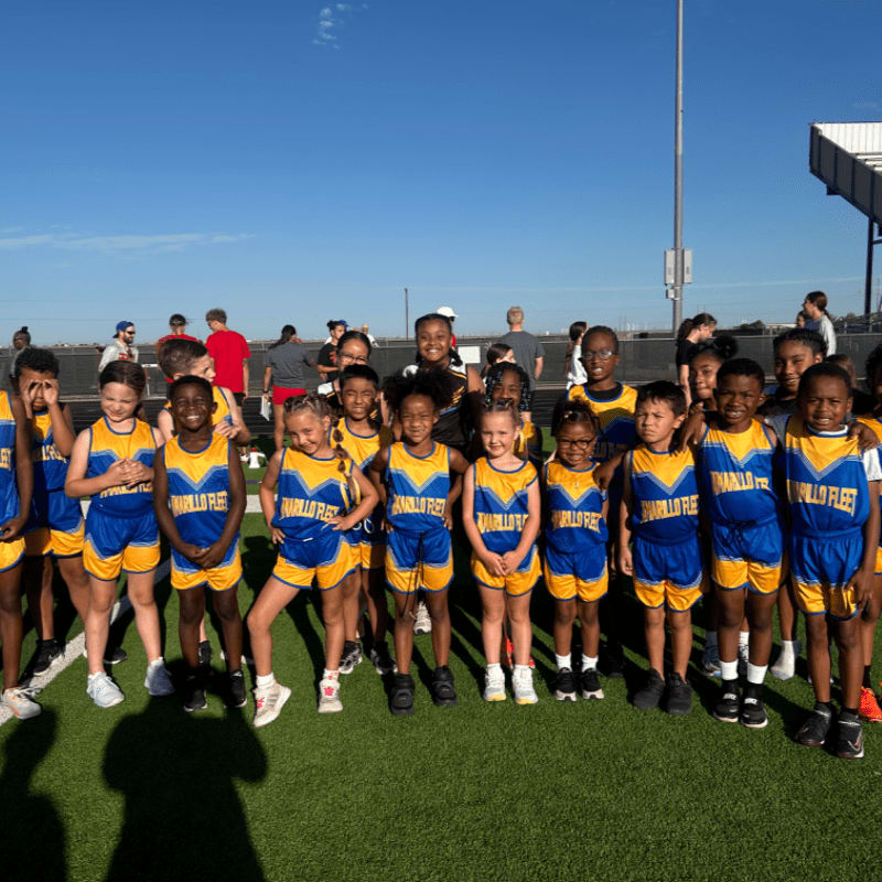 A group of young athletes in blue and yellow uniforms stand on a green track with a stadium and clear blue sky in the background.