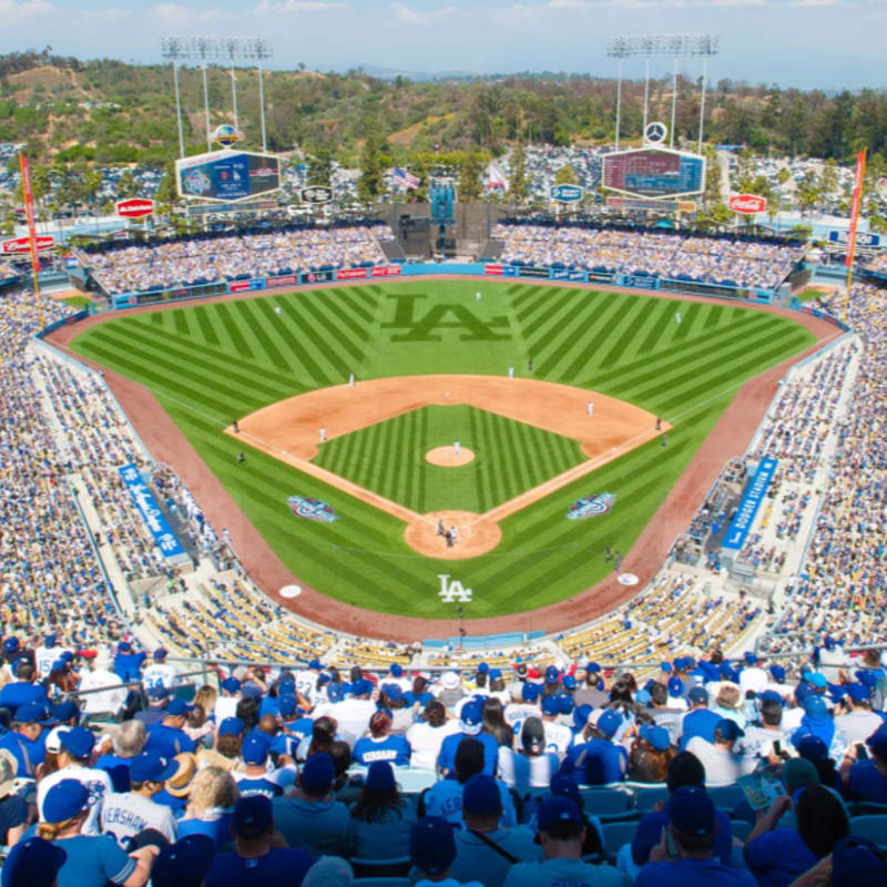 A baseball game is in progress on a sunny day at Dodger Stadium, with a packed crowd in the foreground and rolling green hills in the background.