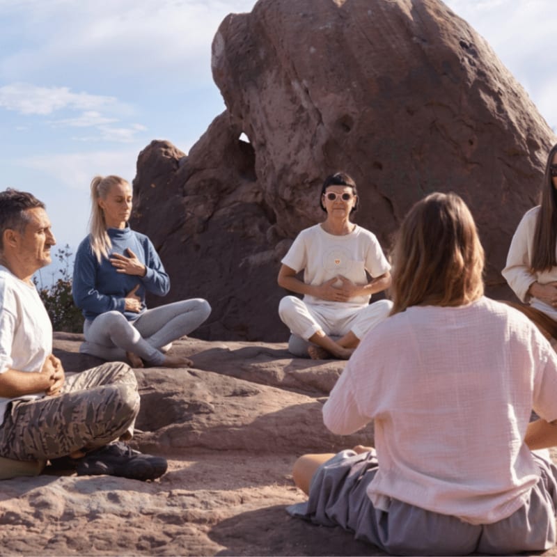 A group of people meditate in a circle on a rocky outcrop with a large boulder in the background.