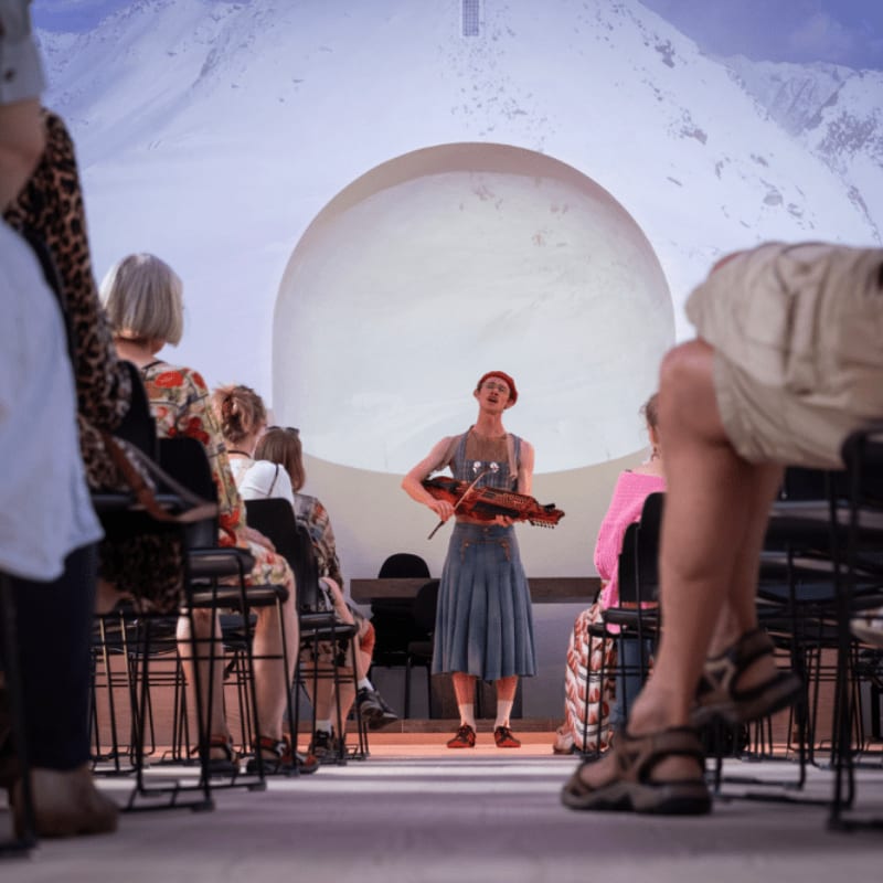 A performer with a violin stands before an audience in front of a large projection of a planet and snowy mountains.