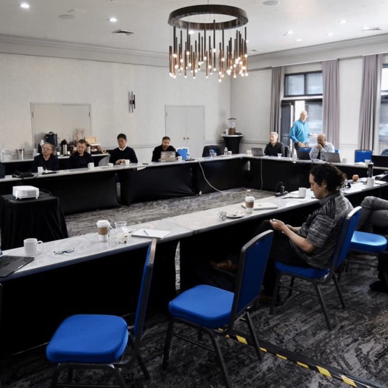 A group of people sit around a U-shaped table in a conference room with a modern chandelier overhead.