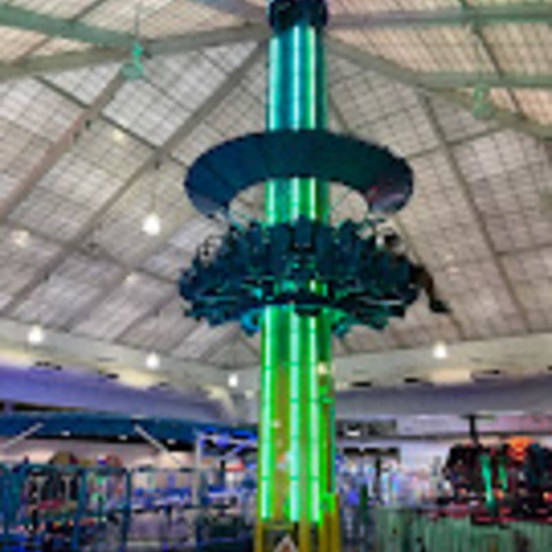 A tall, green-lit amusement park drop tower with a circular seating platform is suspended from a domed ceiling with various arcade games visible in the background.
