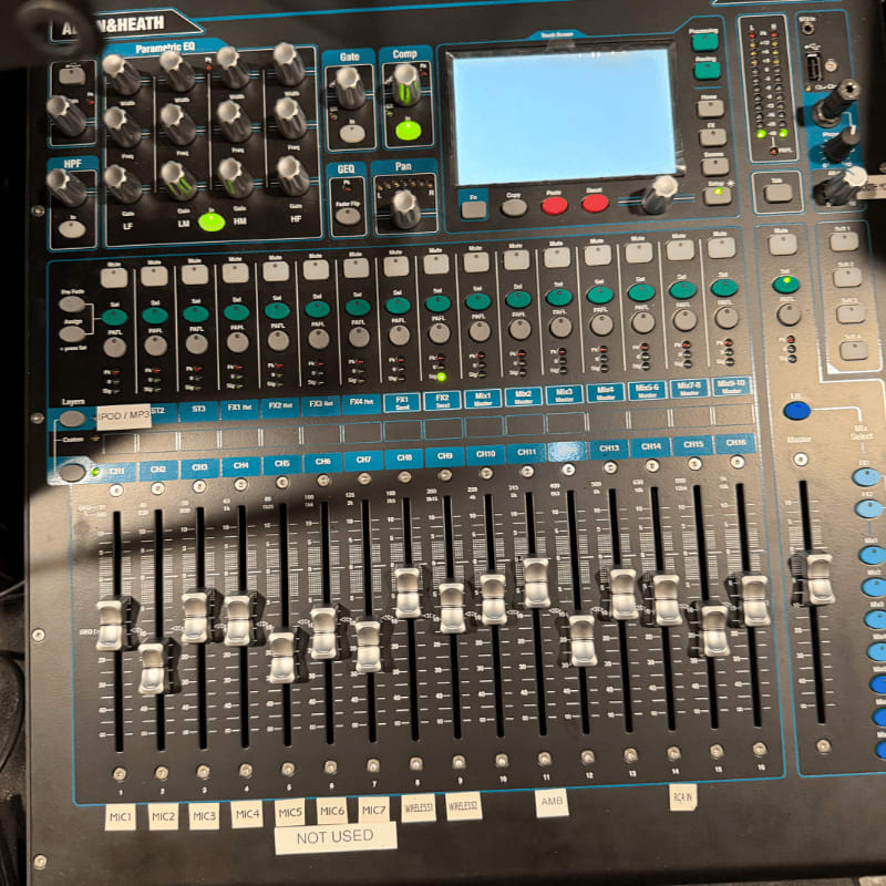 A close-up view of a digital audio mixer with numerous faders and knobs in the foreground, and a bright screen displaying a blue interface in the background.
