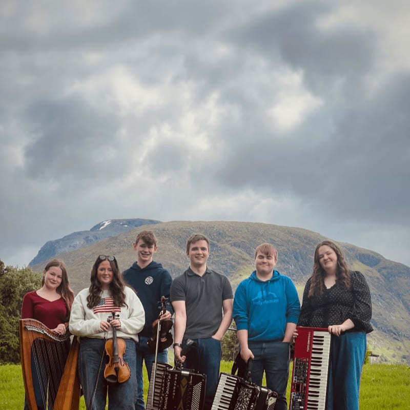 Six young musicians, holding instruments like a harp, violin, and accordions, stand in a grassy field with a large, cloudy mountain in the background.
