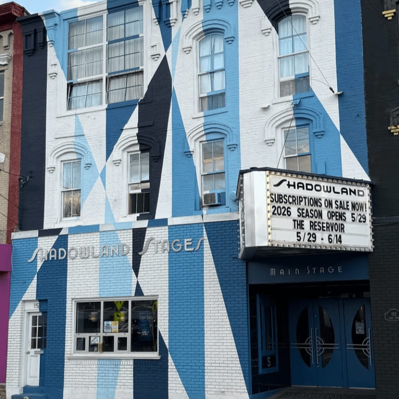 A white brick building with a blue diamond pattern facade features a marquee announcing "SHADOWLAND STAGE" and upcoming shows in the foreground, while the background shows the upper floors of the building with windows.