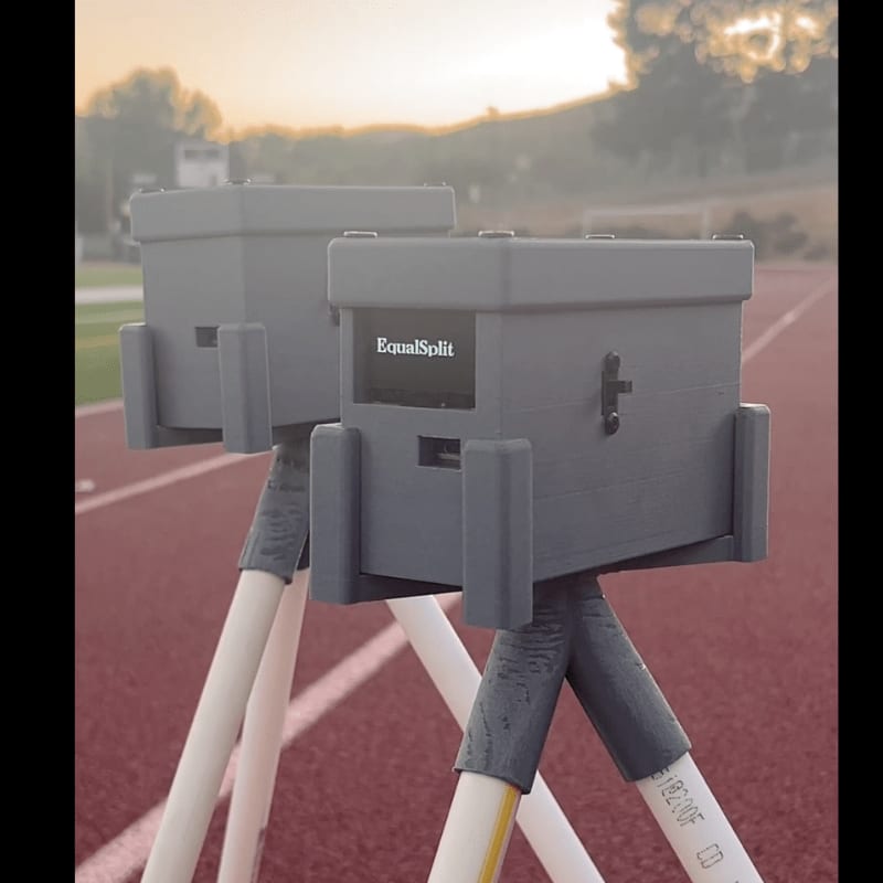Two gray timing devices with "EqualSplit" displayed on their screens stand on tripods on a red track, with a hazy, tree-lined background under a soft sky.