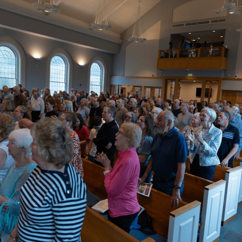 A congregation stands and applauds in a church with arched windows and a balcony in the background.