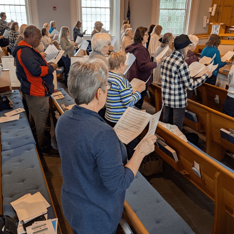 A choir of people in a church, with rows of blue pews in the foreground and large windows in the background.