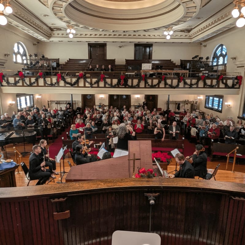 A string orchestra performs for a seated audience in a decorated hall with balconies.