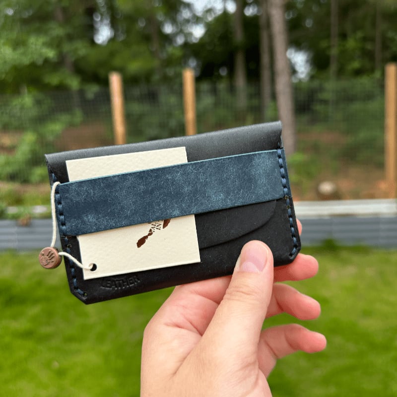A hand holds a dark leather wallet with a blue strap and tags in front of a blurred background of trees and fencing.