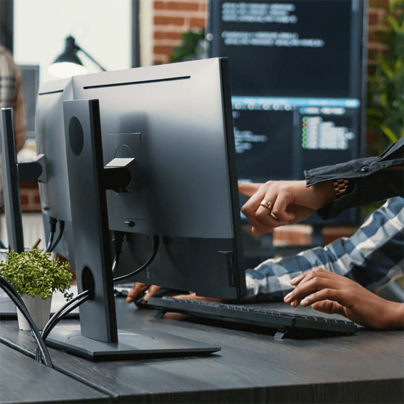 Hands type on a keyboard in front of a computer monitor displaying code, with a brick wall and another monitor in the background.