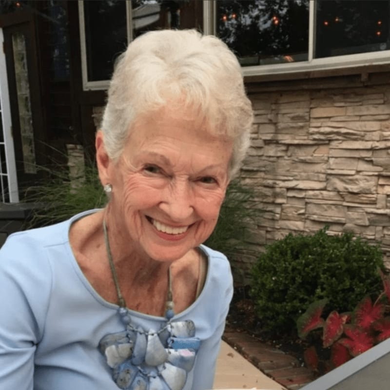 A smiling elderly woman with white hair and a blue top is in the foreground, with a stone wall and greenery in the background.