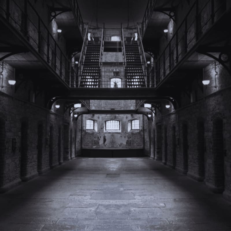 A stark, empty prison cell block with a central walkway leading to a grand staircase in the background.