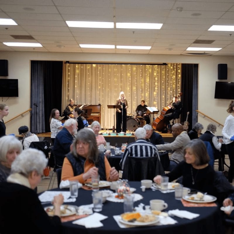 In the foreground, people are seated at round tables with food, while in the background, a band performs on a stage with a shimmering curtain.