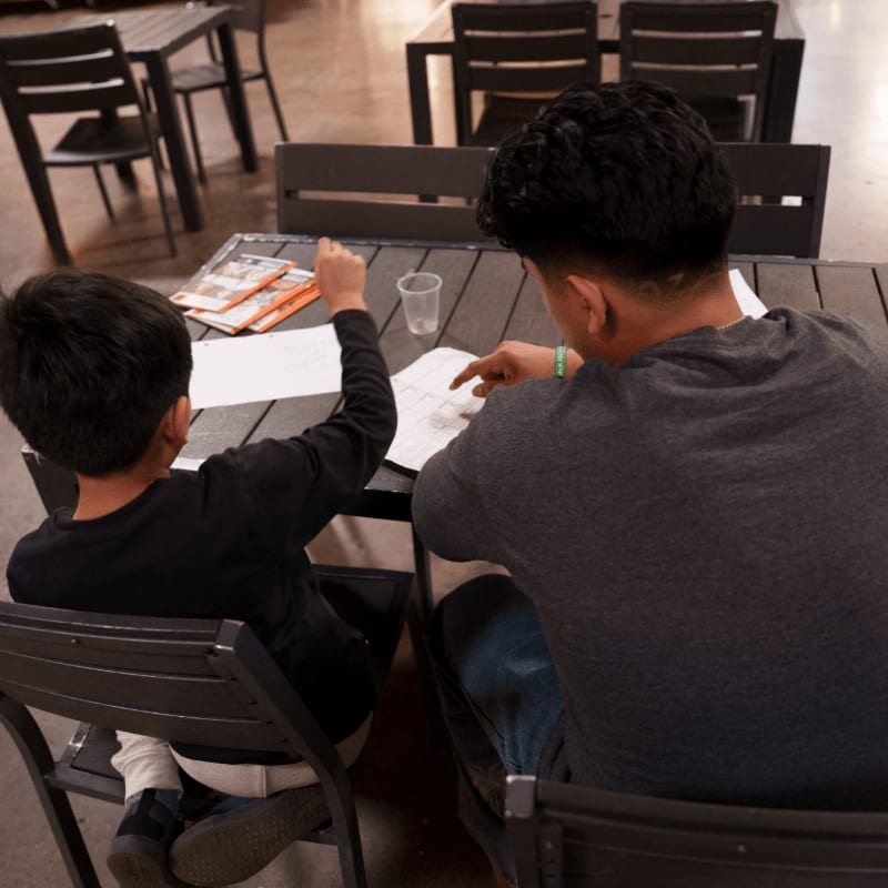 A young boy and an adult sit at a table with papers and a cup, while blurred tables and chairs fill the background.