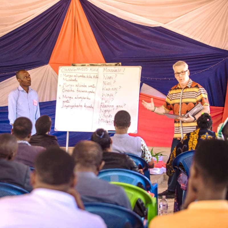 A man in a colorful shirt gestures towards a whiteboard with writing on it, while another man stands to his left, as a seated audience watches in the foreground, all under a draped red, white, and blue fabric canopy.