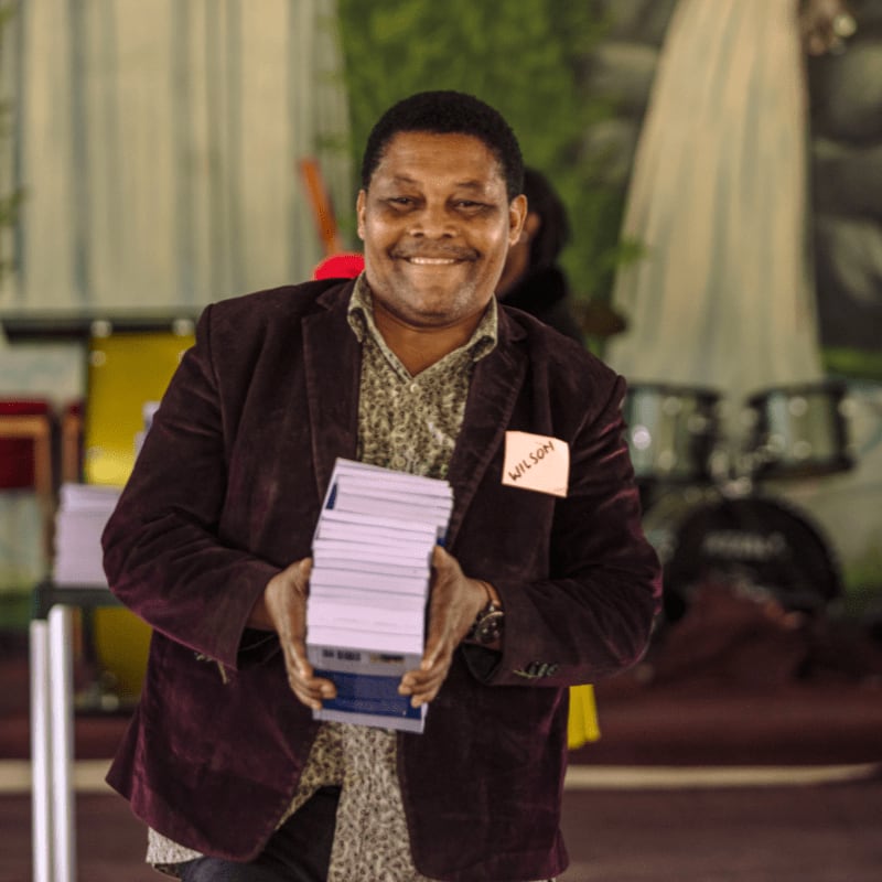 A smiling man in a maroon jacket holds a stack of books in the foreground, while a blurred background reveals a stage with musical instruments and people.