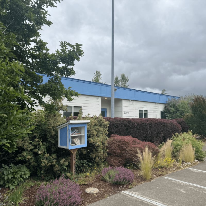 A small blue book-sharing box stands in the foreground amidst lush greenery, with a white building with a blue roof and a cloudy sky in the background.